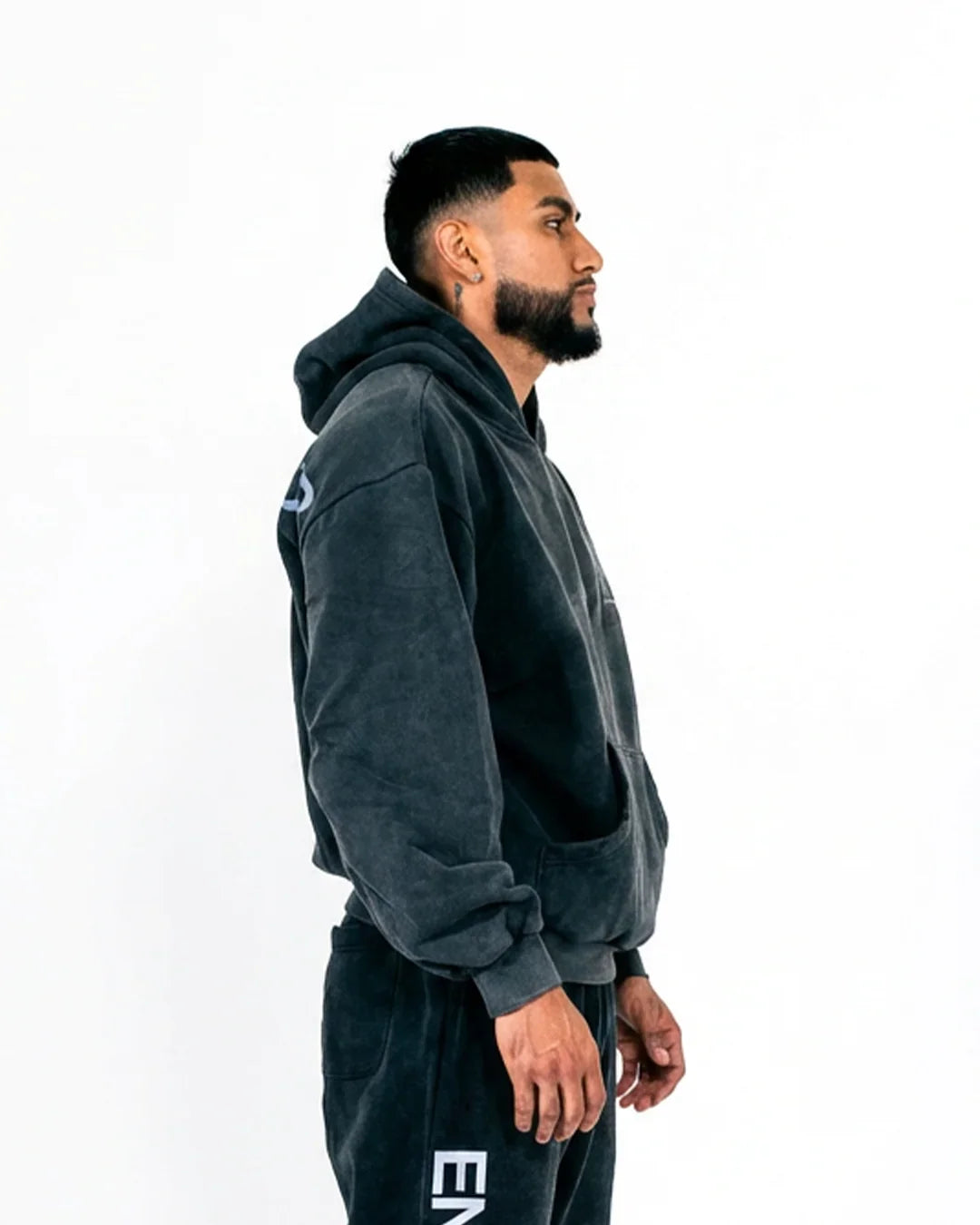 Side profile of man wearing dark charcoal hoodie and matching sweatpants with white lettering on pants against white background