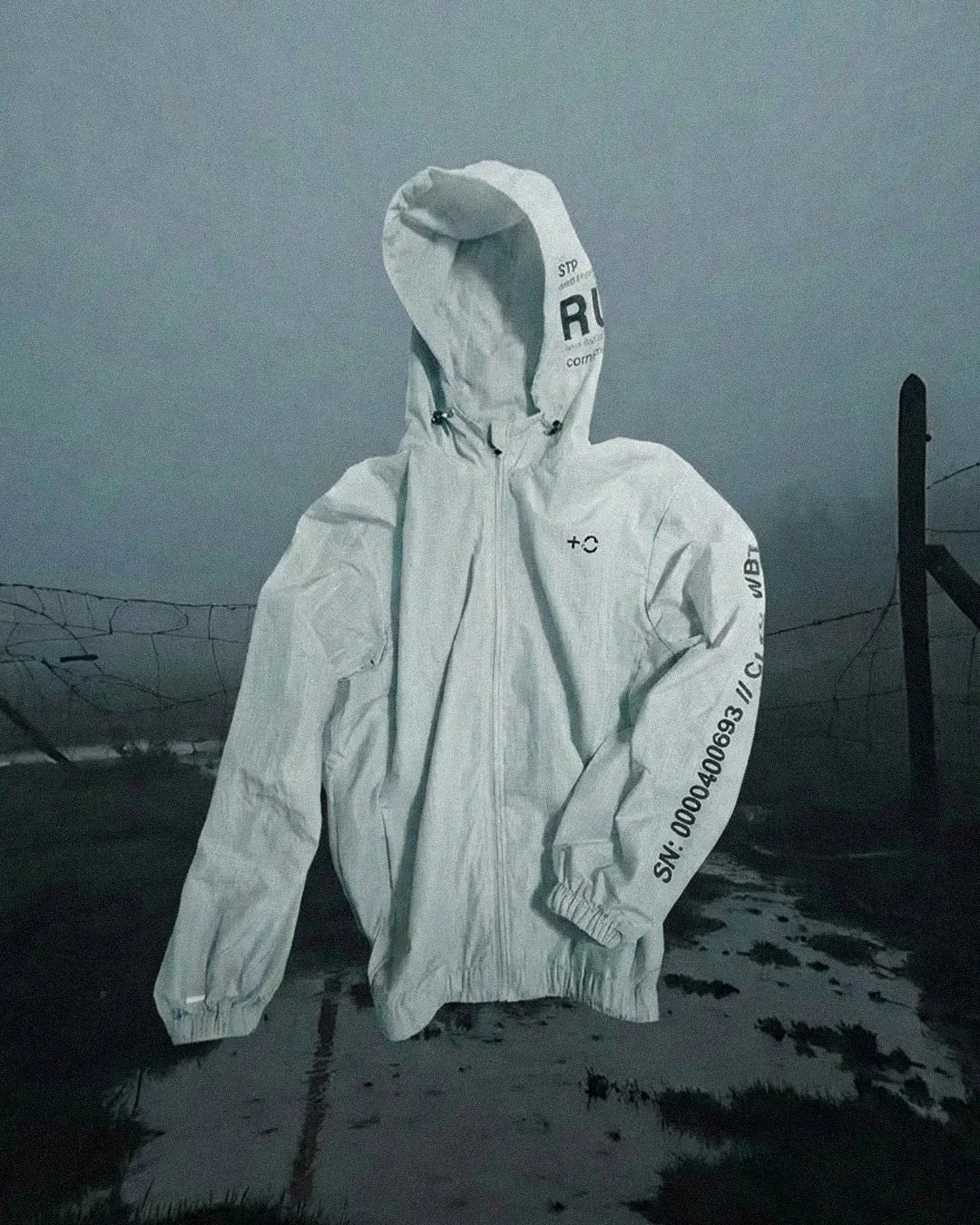 White hooded jacket with black text and serial number on sleeve, floating over misty outdoor scene