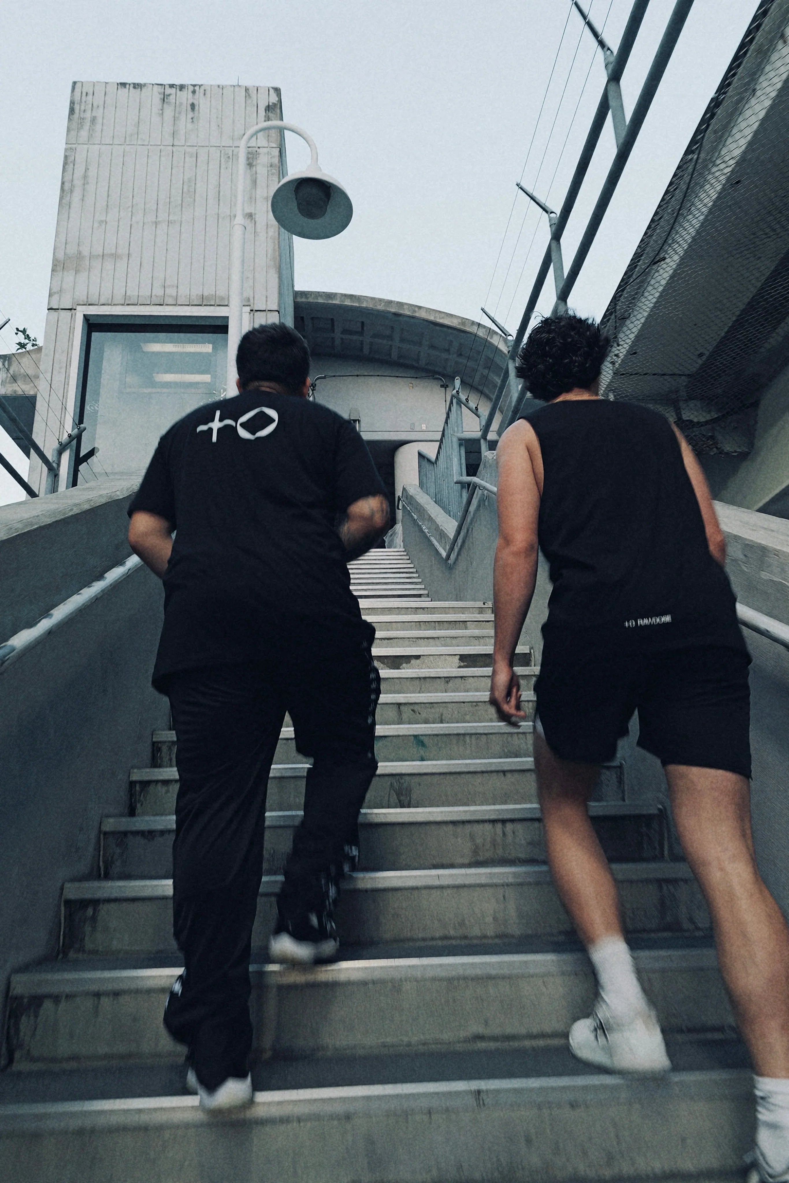 Two men in black sportswear running up concrete stairs outdoors under cloudy sky