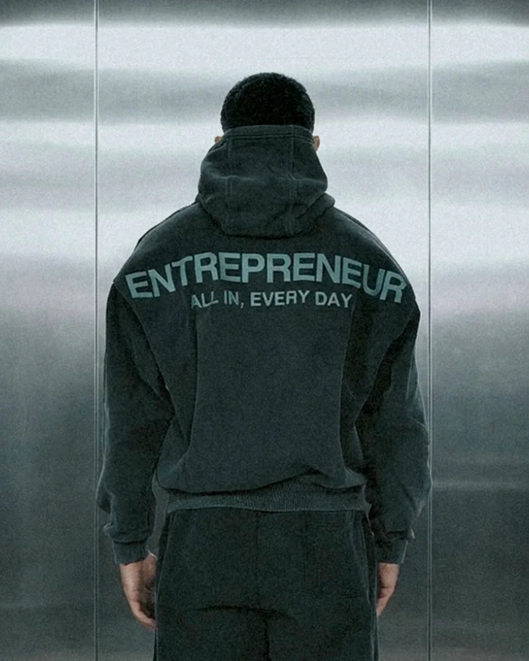 Person wearing dark hoodie with ENTREPRENEUR and ALL IN, EVERY DAY text on back in minimalist setting